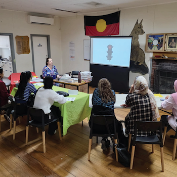 A road rules information session in progress beneath an Aboriginal flag, with participants seated around tables facing a projected traffic light image.